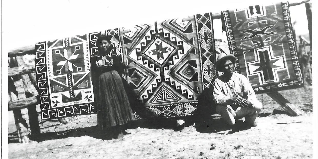 Two women in traditional attire pose by a large patterned carpet.